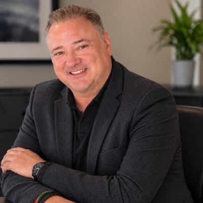 smiling man in a charcoal suit coat and black dress shirt sitting at a boardroom table.