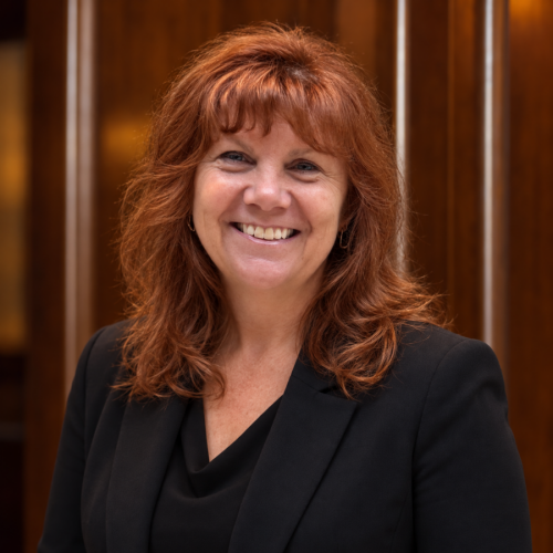 portrait of a smiling woman with red hair and black blouse against a dark wood background